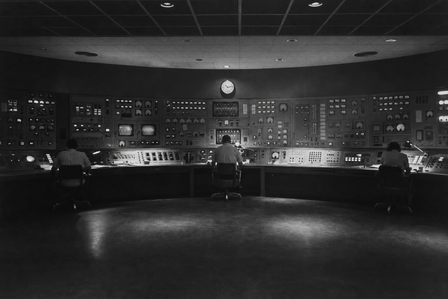 Operators sit before a long wall of analog instruments in a dim institutional control room