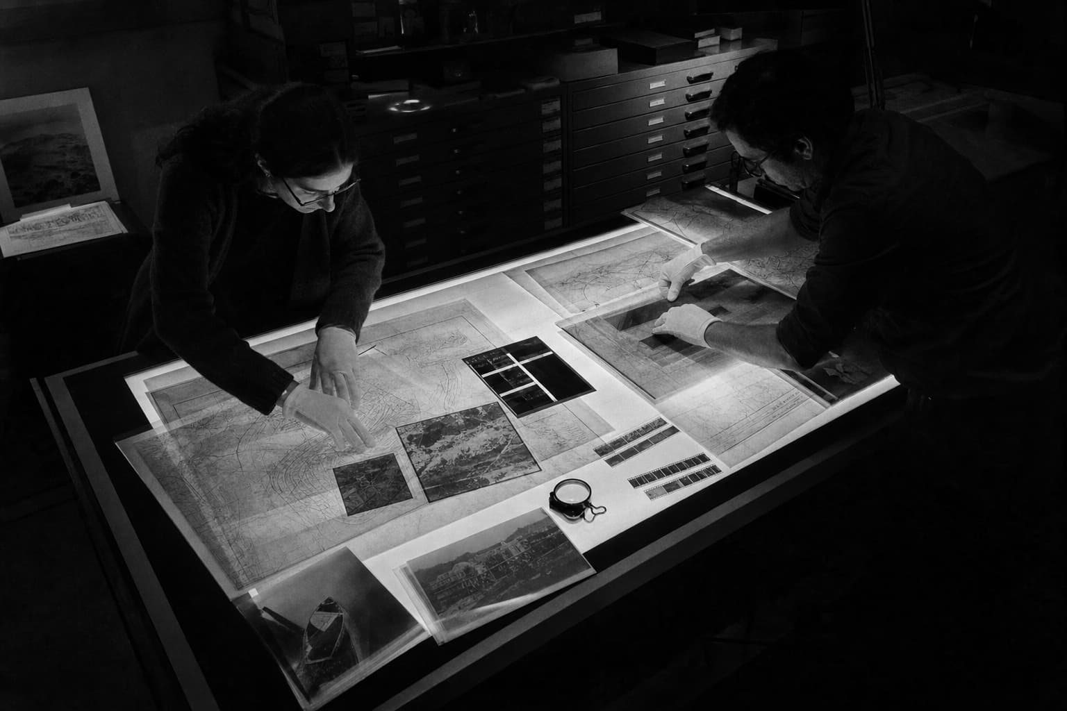 Two archivists arranging maps and photographs across a worktable